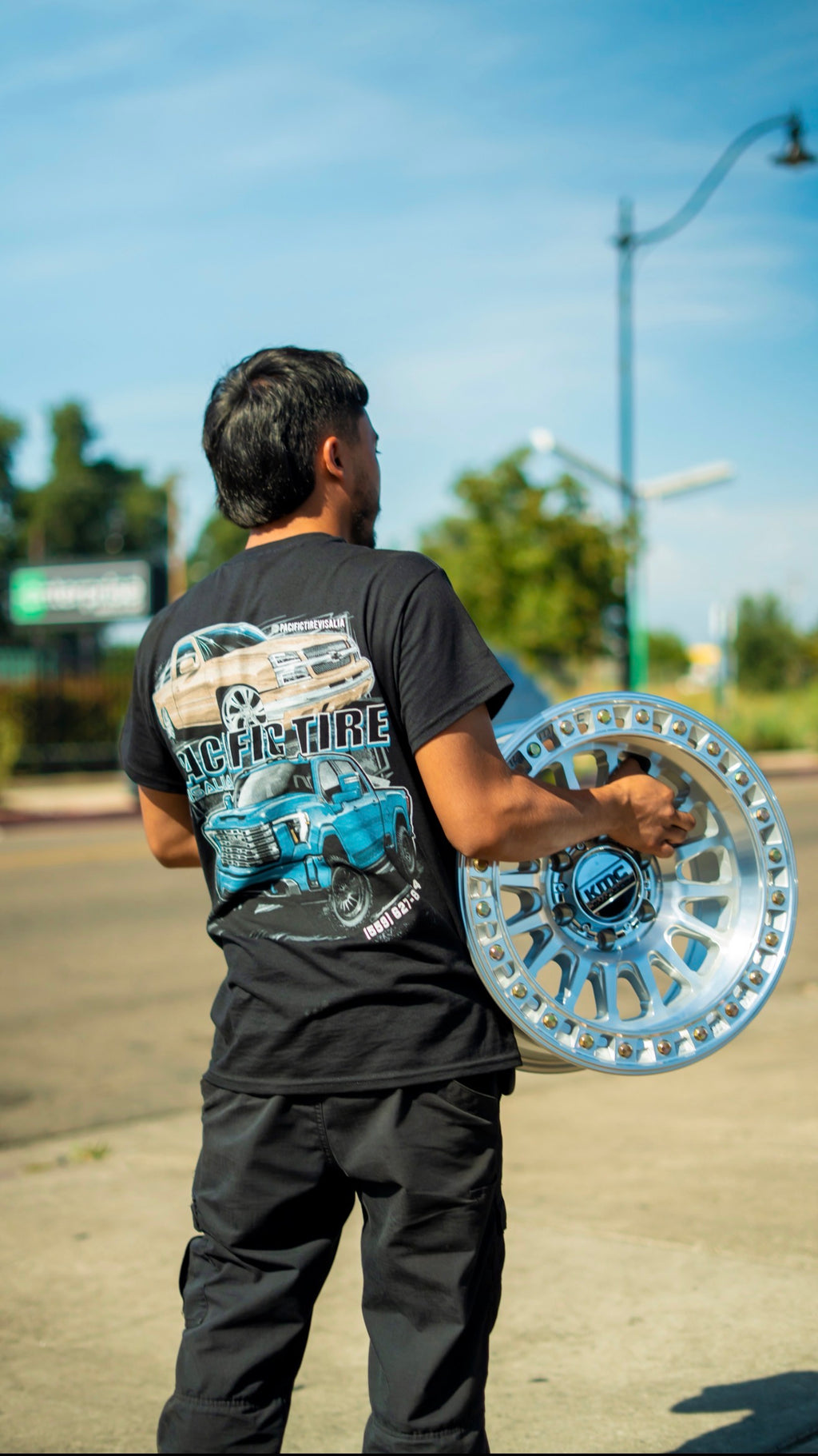 Pacific Tire Visalia truck tee worn outside the shop holding a KMC wheel